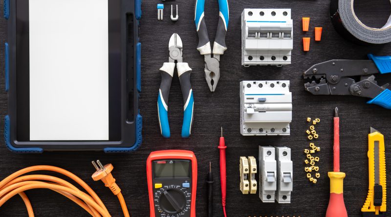 Zestaw narzędzi i różnorodne art elektryczne, w tym bezpieczniki, złączki, multimetr i przewody, ułożone na ciemnym blacie