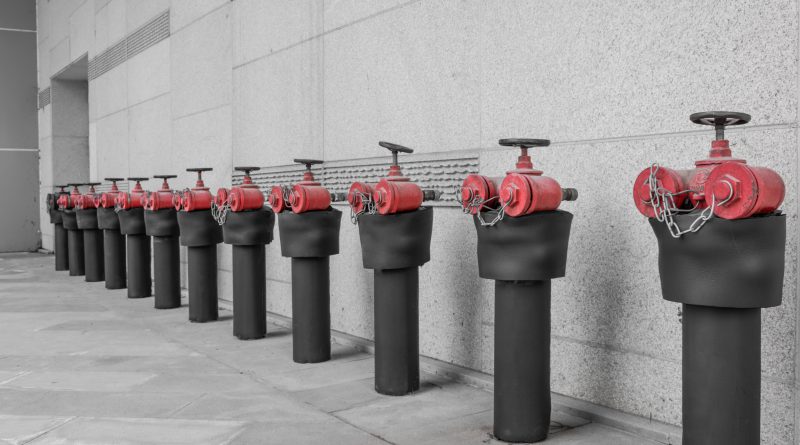 Rząd czerwono-czarnych zaworów hydrantowych przy betonowej ścianie, stanowiących element systemu przeciwpożarowego zasilanego wodą, którą magazynują zbiorniki ppoż naziemne.